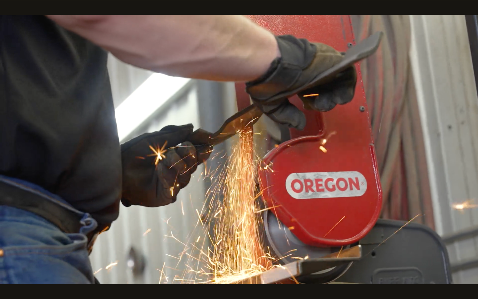 Gloved worker sharpening a mower blade on a red Oregon bench grinder as sparks fly.