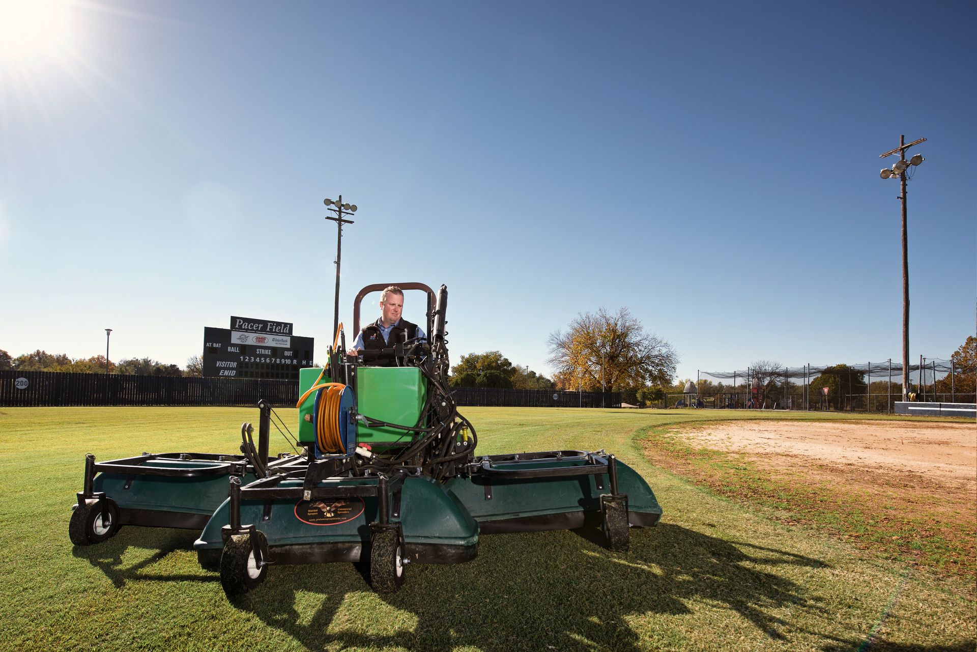 Operator driving a shielded sprayer attachment with tank and hose reel across a ballfield outfield.