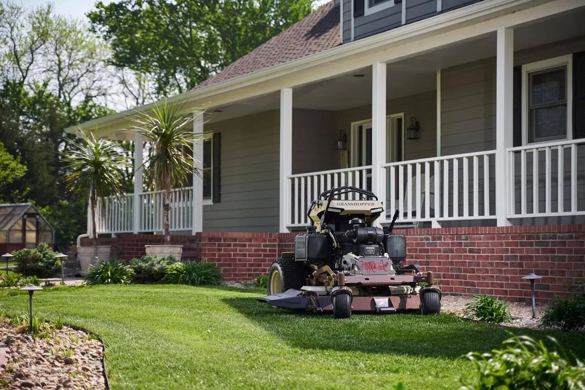 Grasshopper stand-on mower parked on a trimmed lawn before a house's covered front porch.