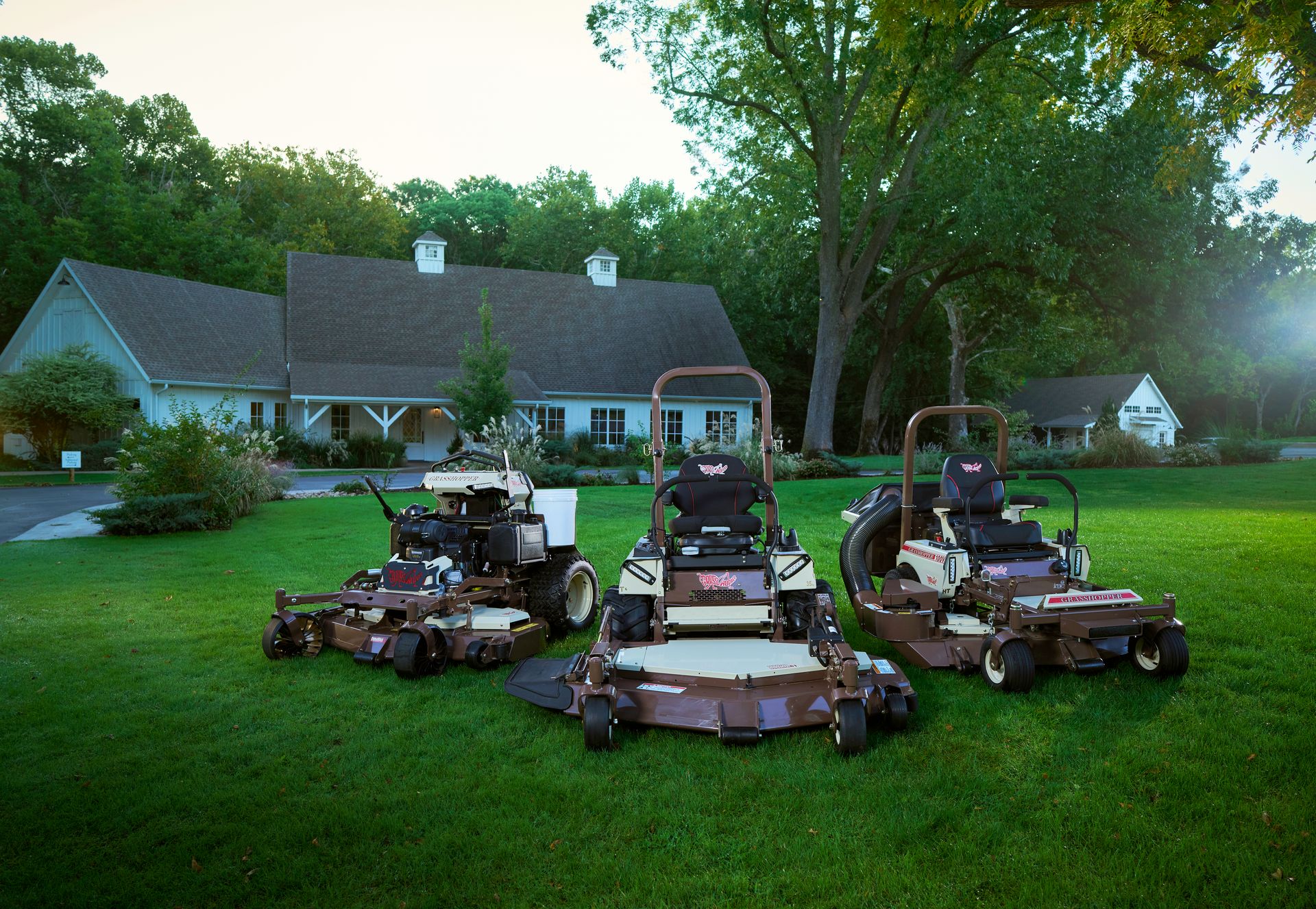 Three Grasshopper mowers, one with a collection hose, parked on lawn before a white barn.