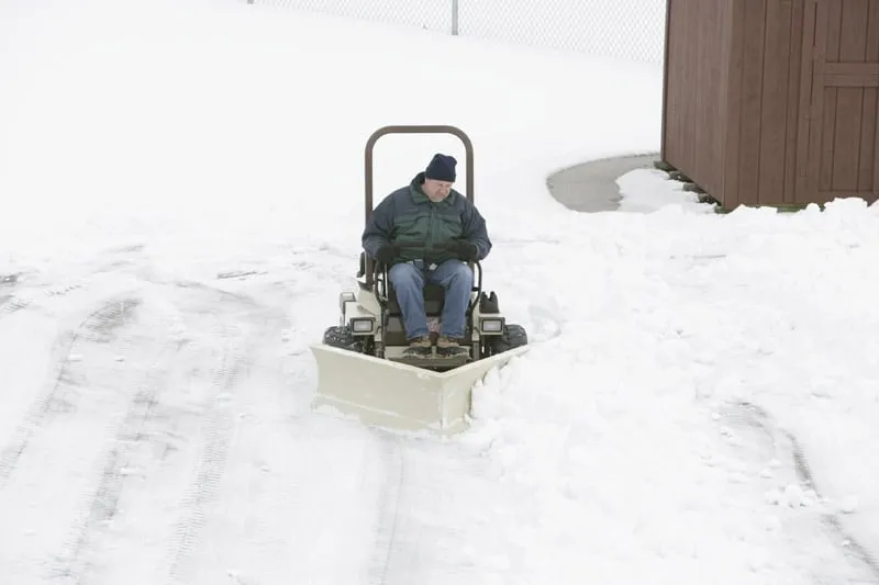 Operator clearing a snowy driveway with a V-plow mounted on a zero-turn machine, shed behind.