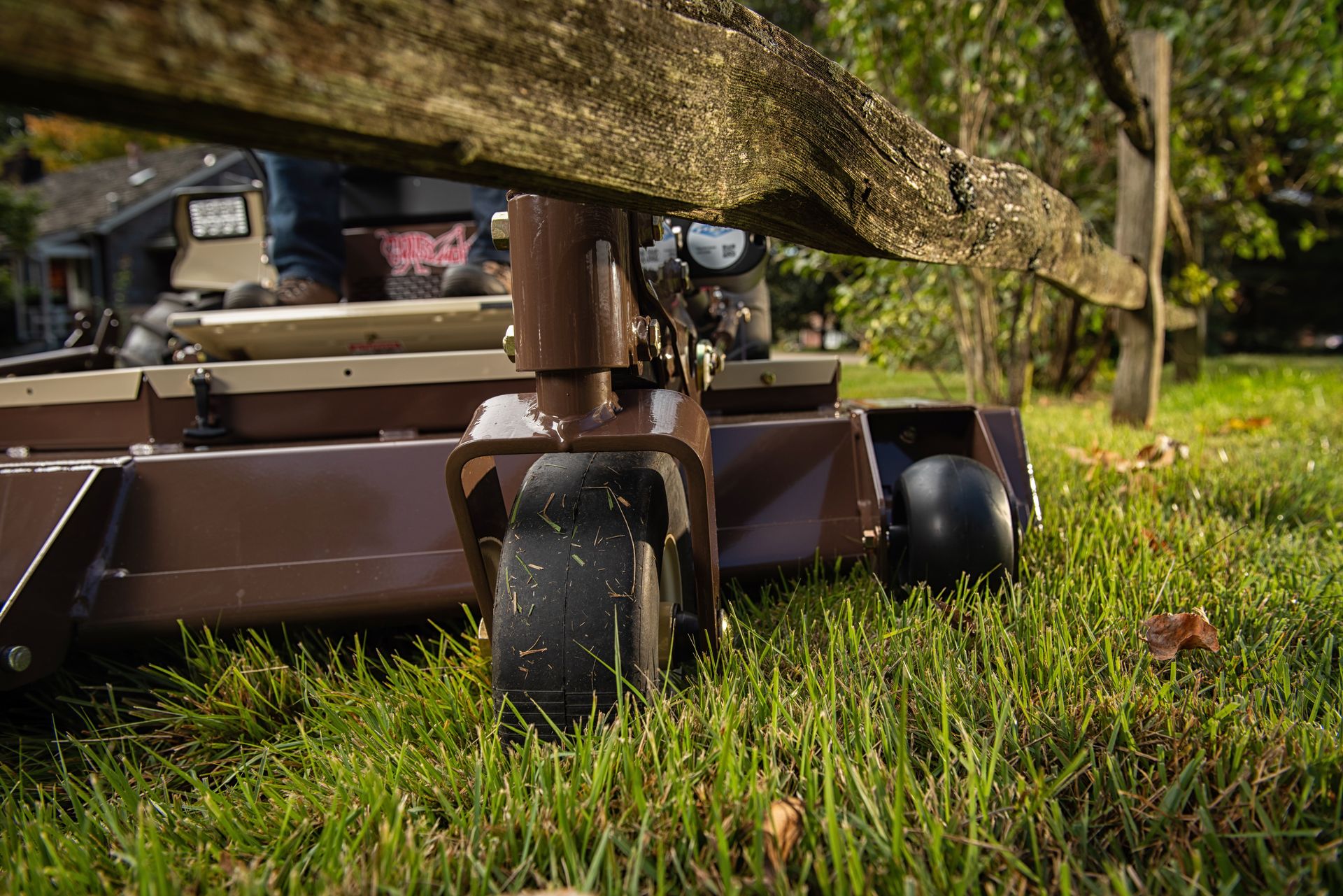 Close-up of a mower's front deck and caster wheel passing beneath a weathered fence rail.