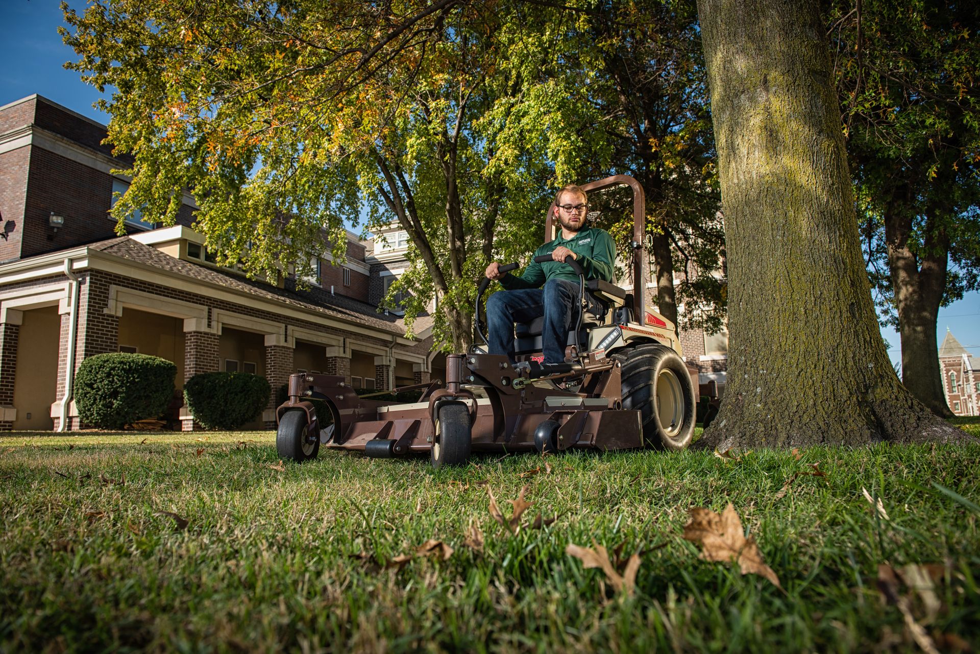 Grounds worker in a green polo mowing under a large tree beside brick campus buildings.