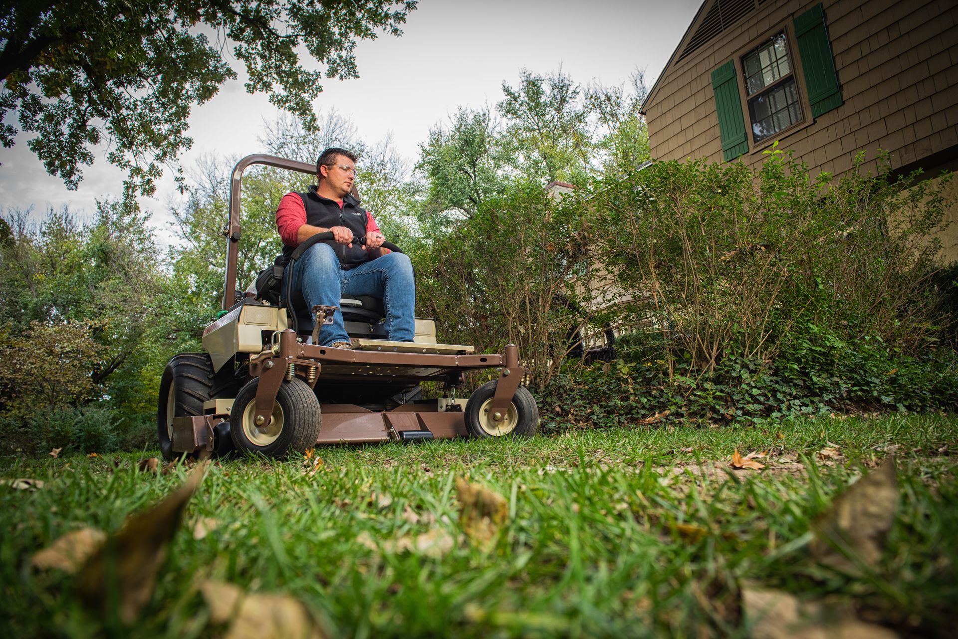 Low-angle view of a man riding a Grasshopper FrontMount mower past overgrown shrubs and leaves.