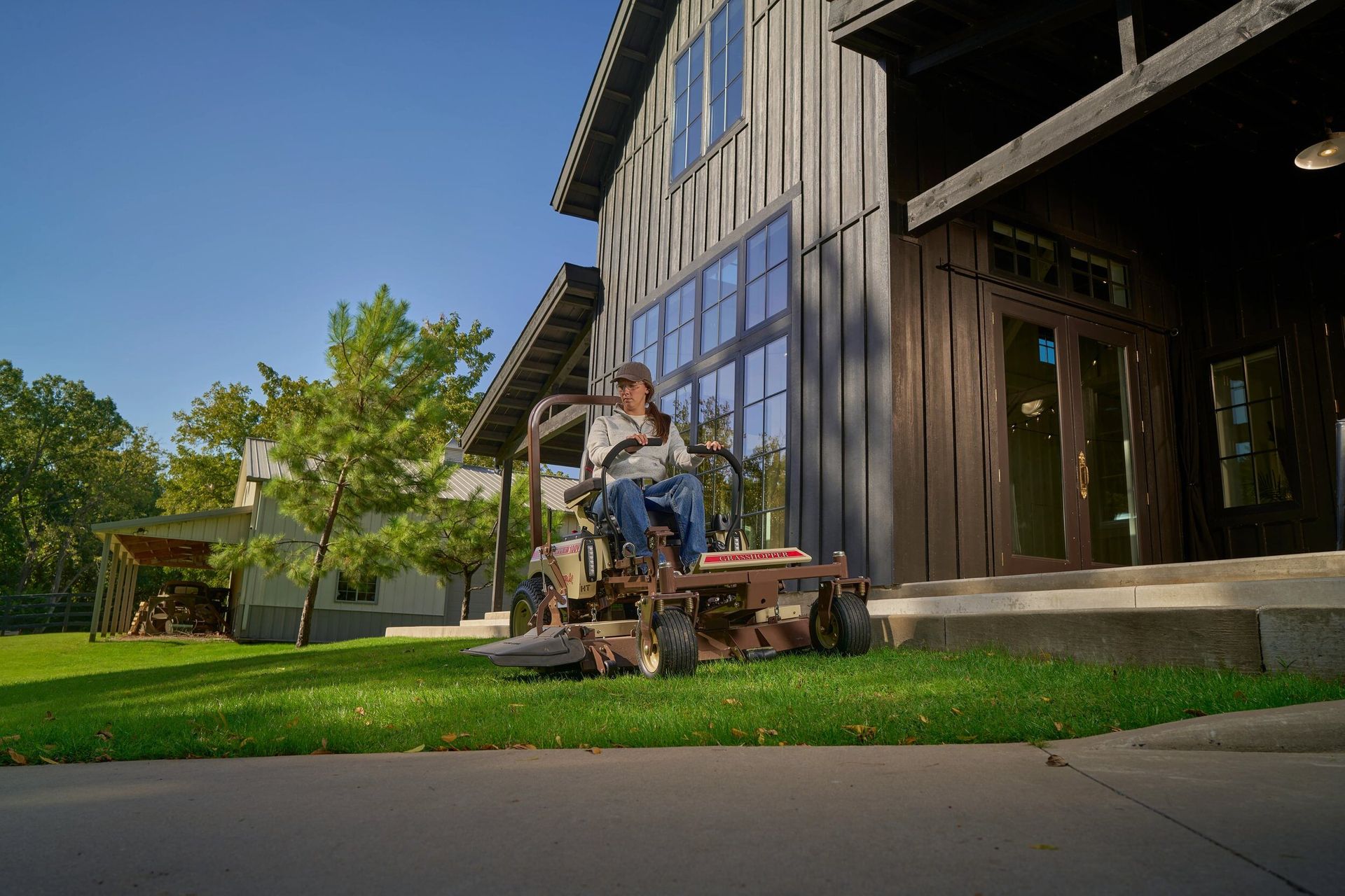 Woman mowing a lawn on a Grasshopper 100V mower beside a modern dark barn-style house.