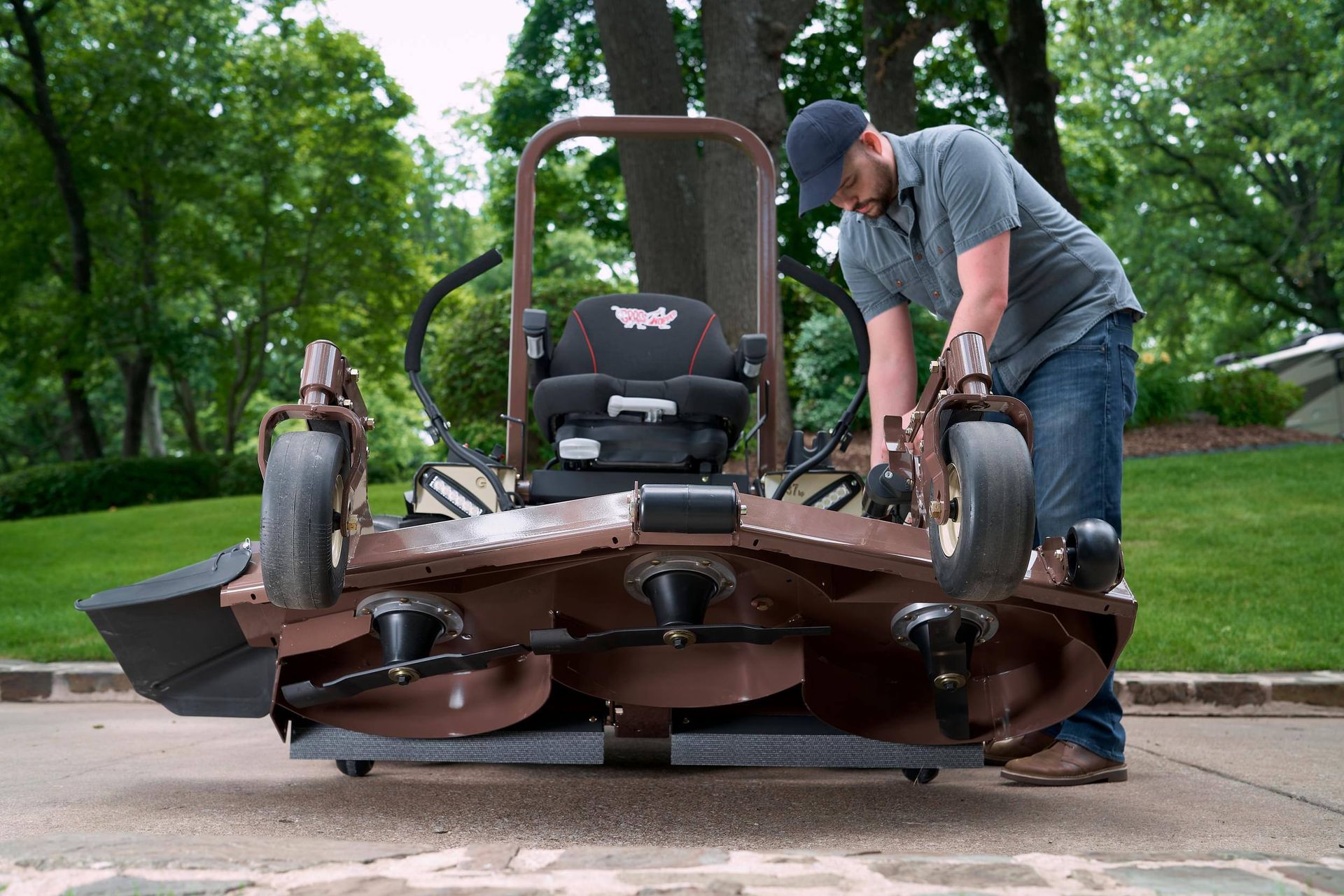 Man tilting up a Grasshopper mower deck on a driveway to expose three blades and spindles.