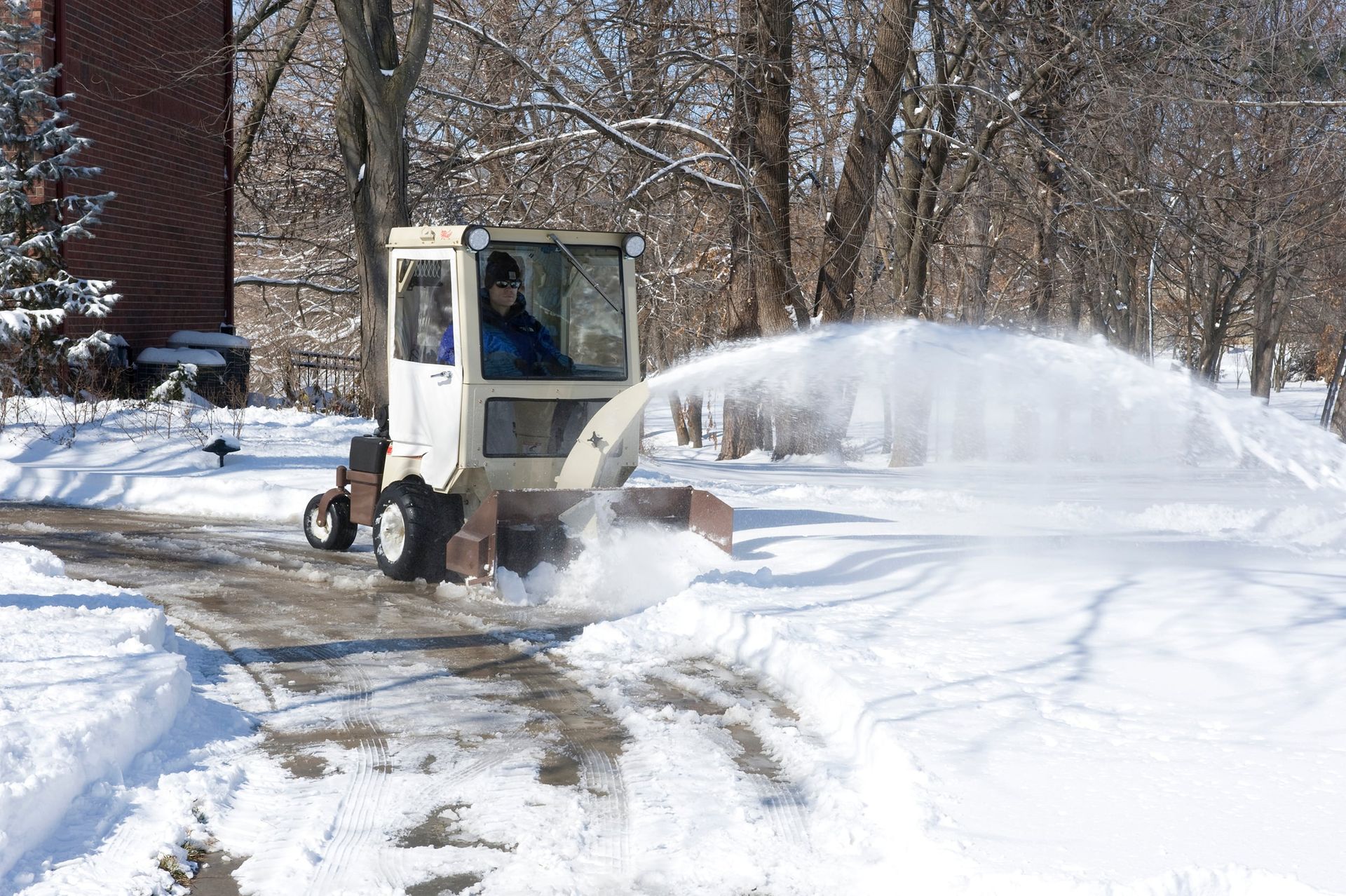 Operator in an enclosed cab clearing a driveway as a snowthrower arcs snow aside.