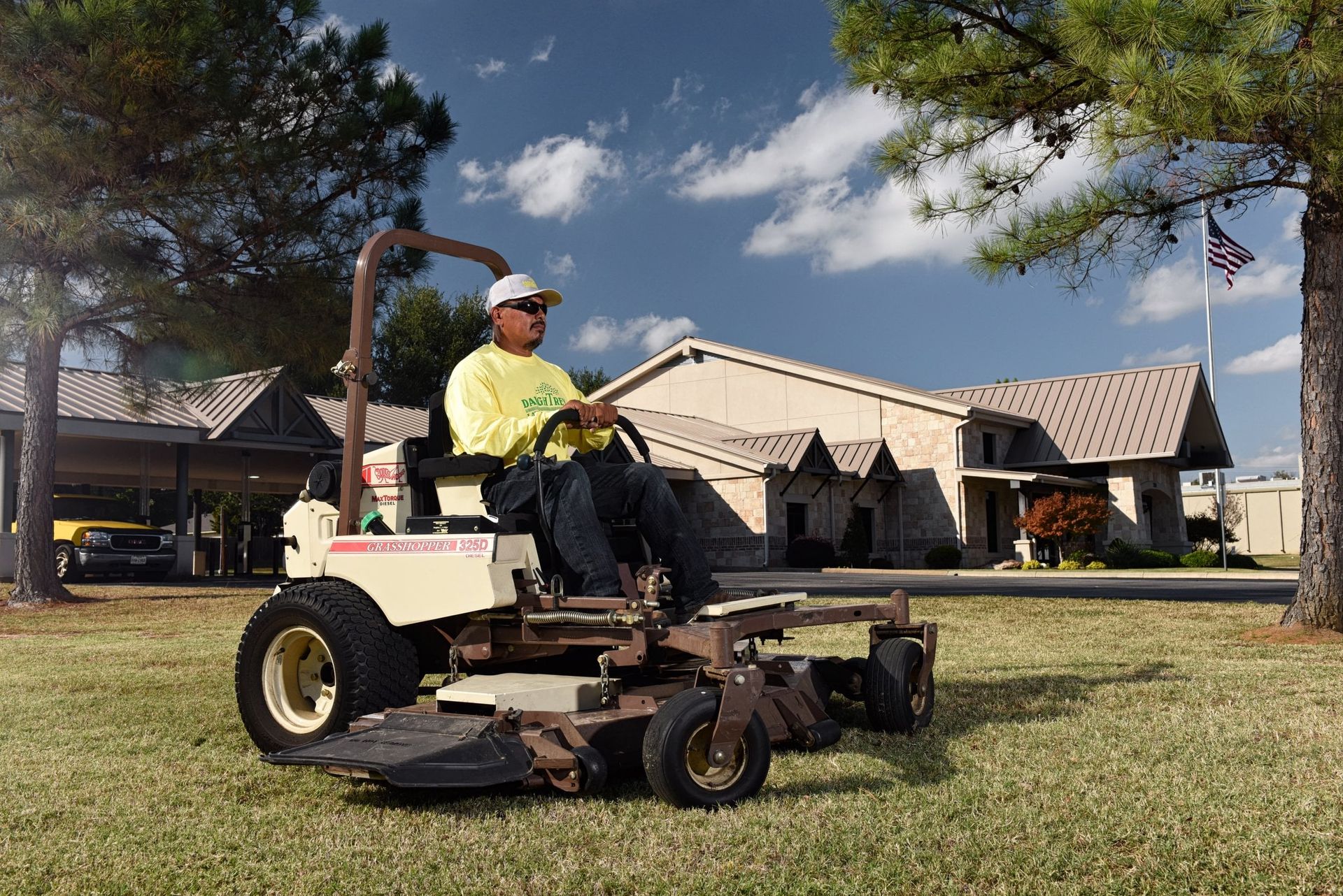 Operator on a Grasshopper 325D diesel FrontMount mower cutting grass outside a stone commercial building.