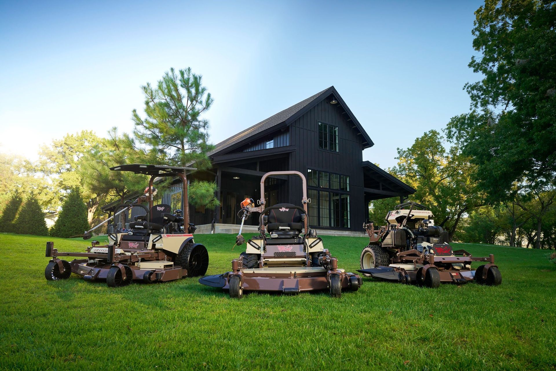 Three Grasshopper mowers parked side by side on a lawn before a black barn-style house.