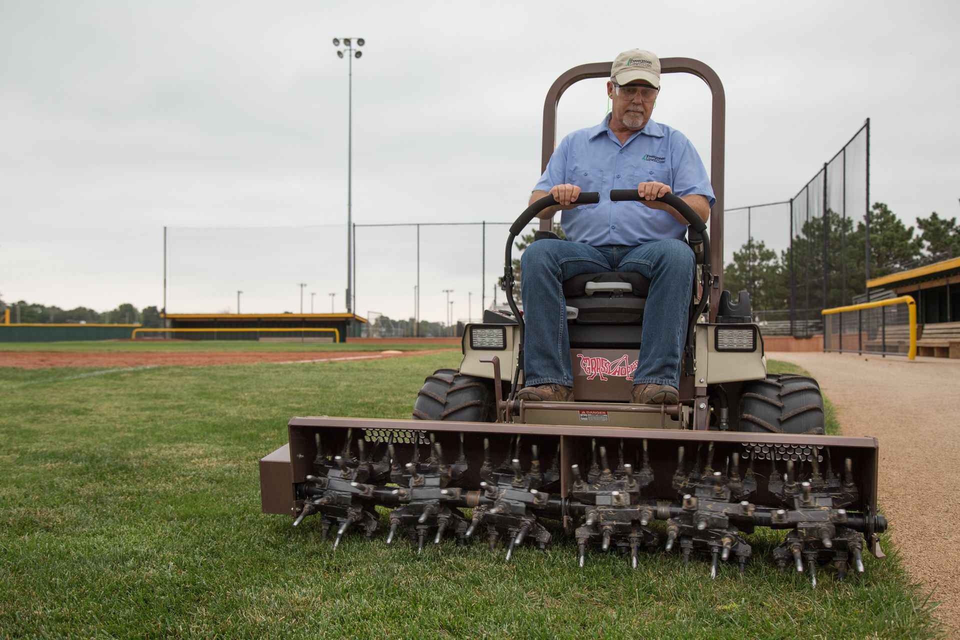 Lawn care operator running a Grasshopper mower's AERA-vator attachment across grass beside a baseball infield.