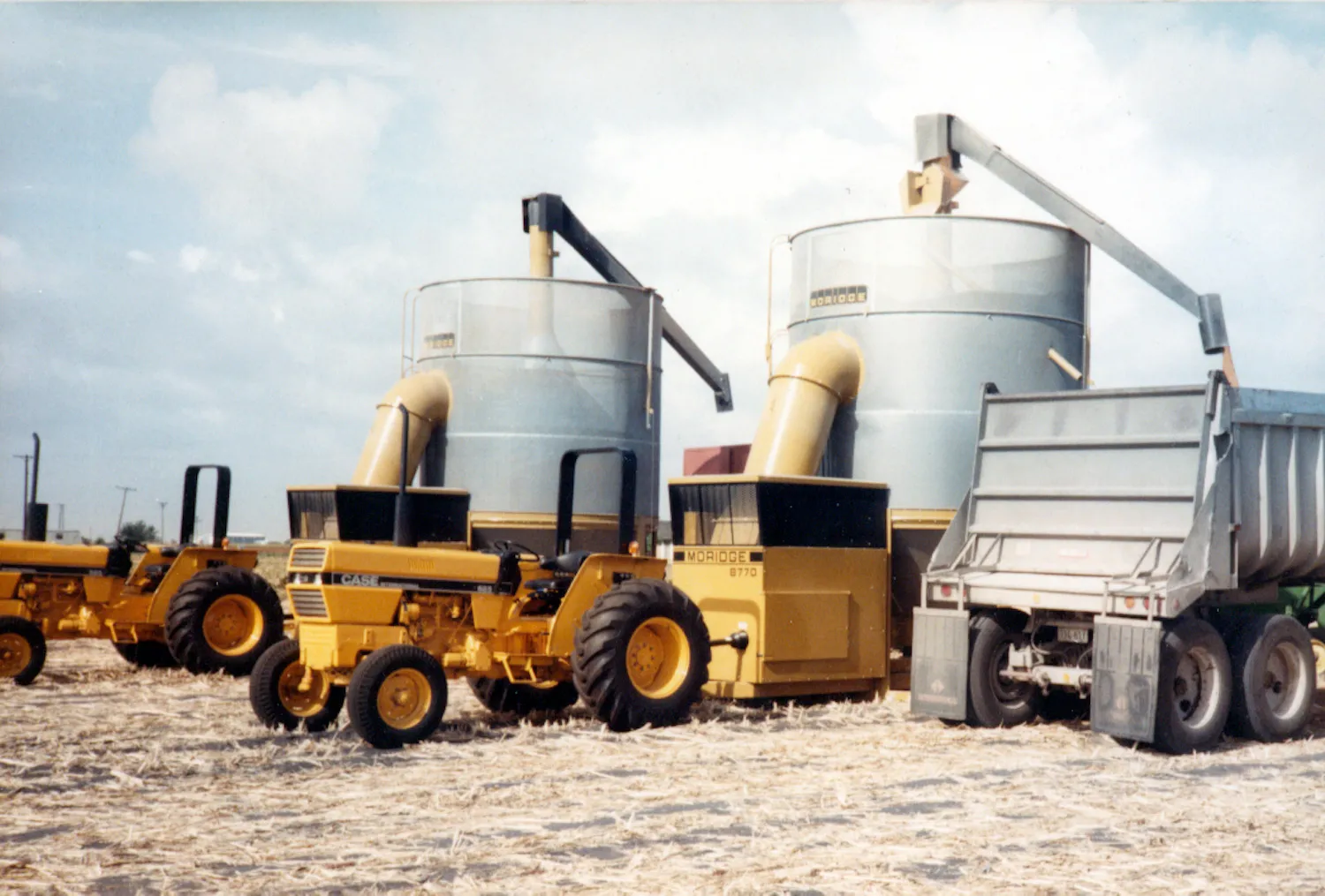 Yellow Moridge grain dryers with augers, a Case tractor, and a dump truck on straw-covered ground.