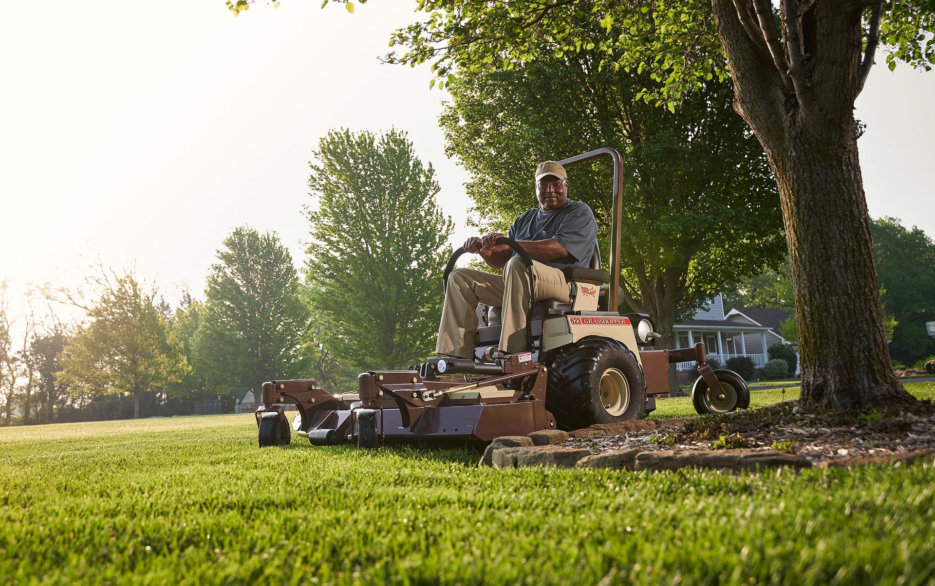 Operator mowing a sunlit residential lawn on a Grasshopper 623 FrontMount mower beside a large tree.