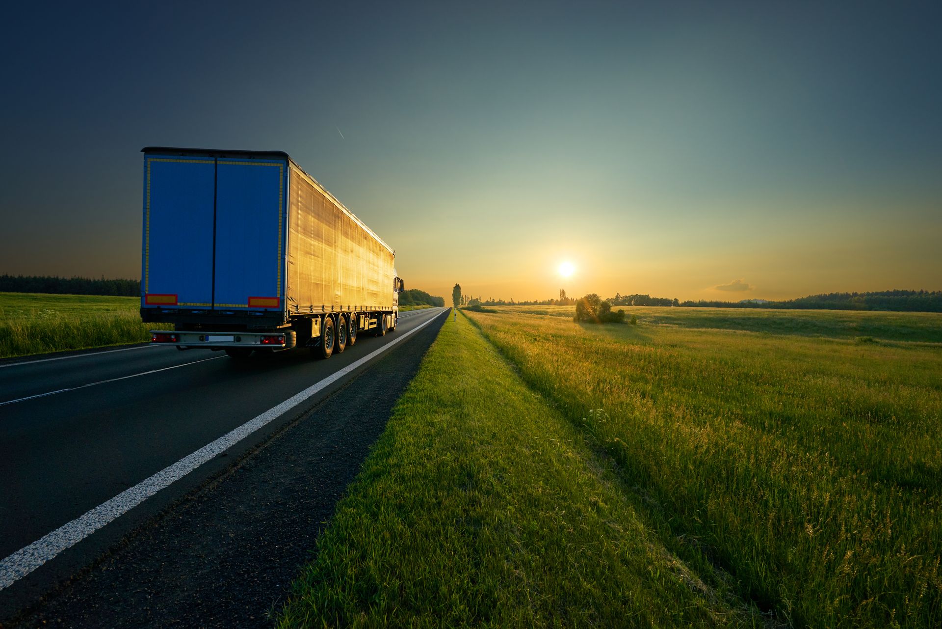 Semi trailer truck on a rural highway at sunset beside a grassy shoulder and open field.