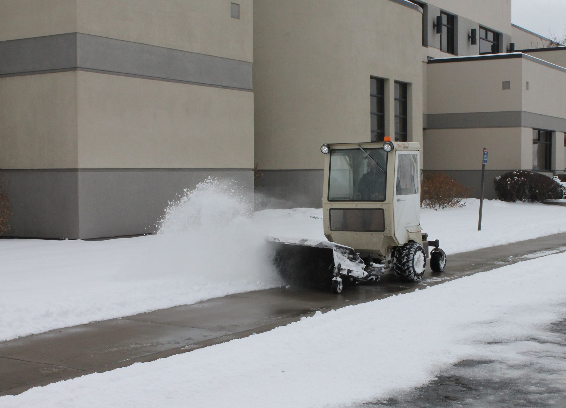 Cab-equipped Grasshopper machine sweeping snow from a sidewalk with a rotary broom attachment.