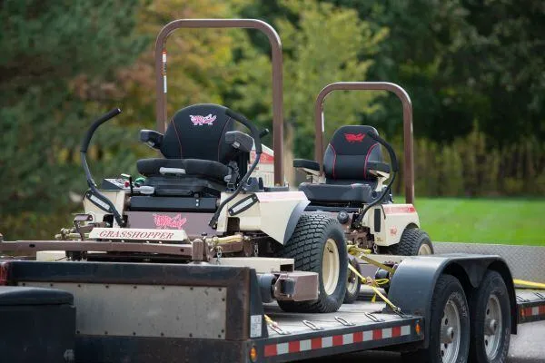 Two Grasshopper mowers with black suspension seats and rollbars loaded on a flatbed trailer.
