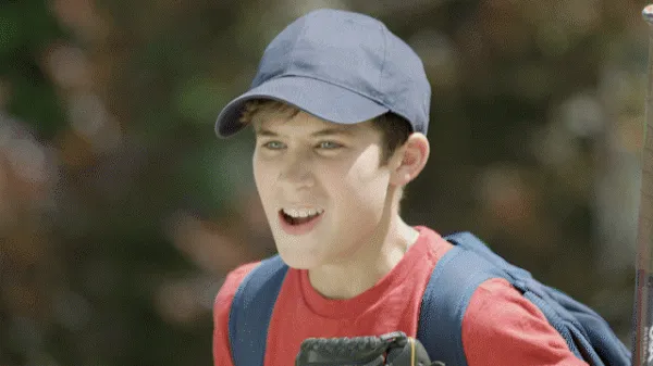 Teenager in a blue ball cap and red shirt talking excitedly outdoors with a backpack.